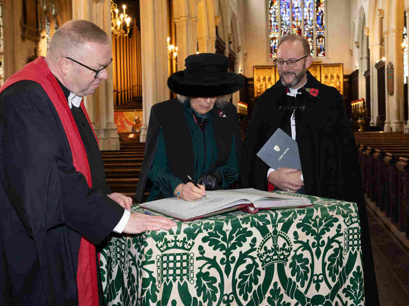 The Queen signs a book as a verger and a priest watch