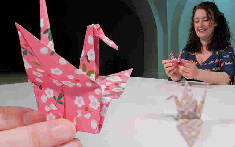Photograph of folded paper crane being held, with a person folding a paper crane in the background