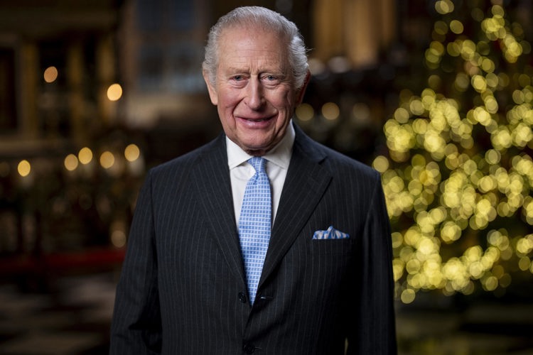 A portrait photograph of HM The King smiling as he stands in the Lady Chapel at Westminster Abbey. A Christmas tree lit with warm white lights stands in the background.