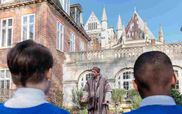 Photograph of adult actor dressed as Peter from the Bible, with the backs of two childrens heads in the foreground and the estate of Westminster Abbey in the background.