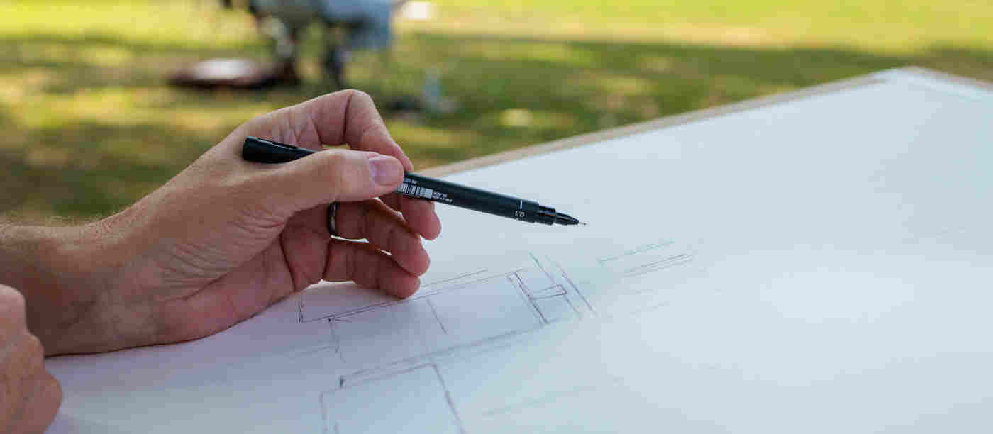 Photograph of a hand holding a pen sketching on a large piece of paper with grass in the background