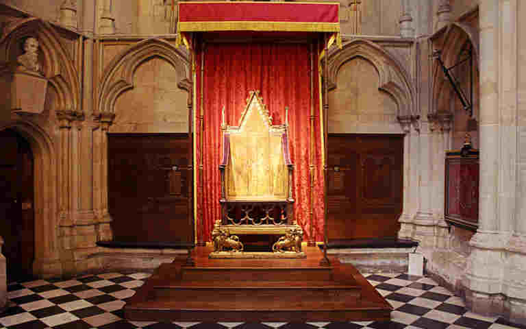 The Coronation Chair on a stepped wooden podium surrounded by gold-fringed red curtain in St George's Chapel, Westminster Abbey