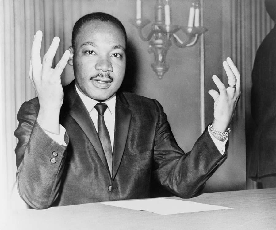 Rev. Dr Martin Luther King Jr seated at a table with a piece of paper in front of him and his hands in the air
