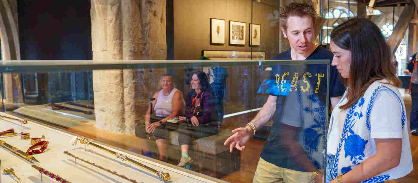Photograph of people looking at a display case of coronation regalia in the foreground, with two people seated in the background, within the Queen's Diamond Jubilee Galleries of Westminster Abbey