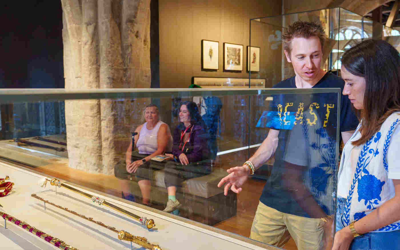 Photograph of people looking at a display case of coronation regalia in the foreground, with two people seated in the background, within the Queen's Diamond Jubilee Galleries of Westminster Abbey
