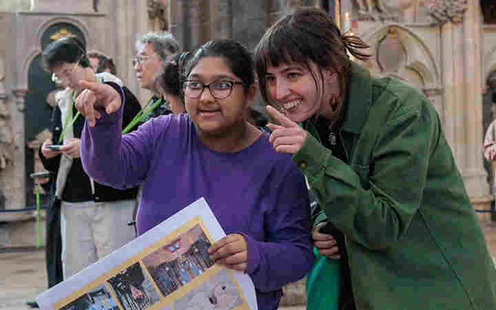 Photograph of young person and accompanying adult smiling and pointing at something within Westminster Abbey, while holding a paper visual story