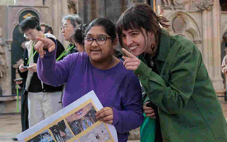 Photograph of young person and accompanying adult smiling and pointing at something within Westminster Abbey, while holding a paper visual story