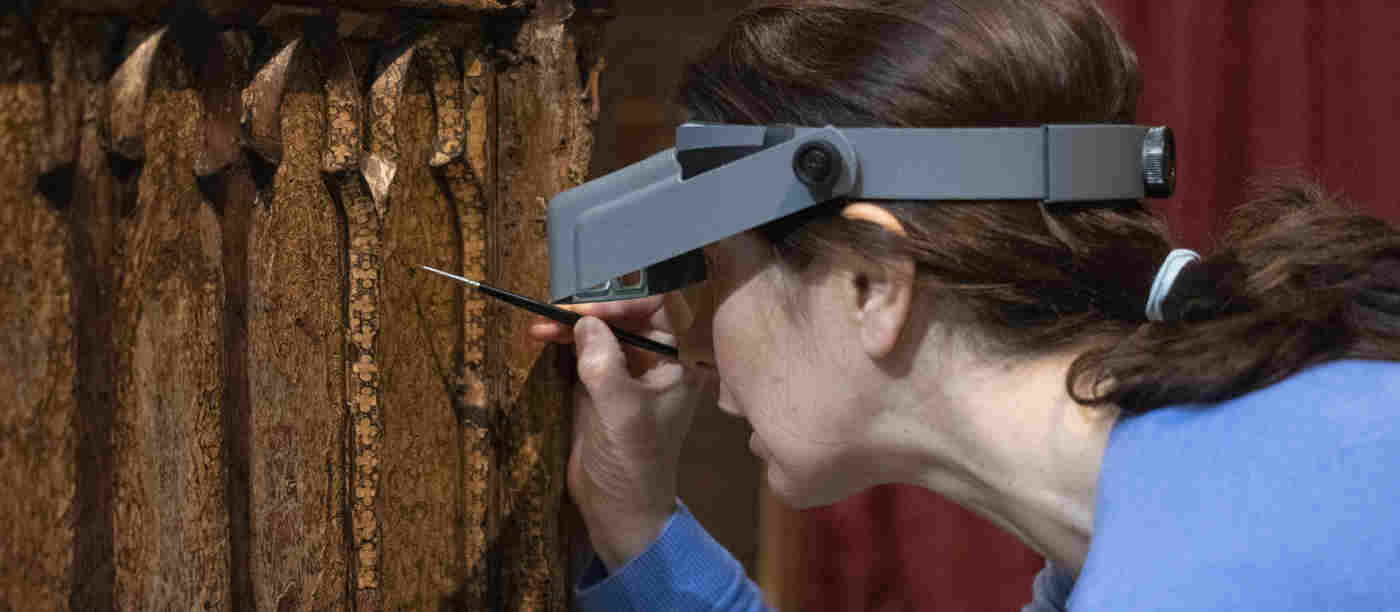 Photograph of woman holding a small paintbrush and wearing a magnifying headset conserving the Coronation Chair