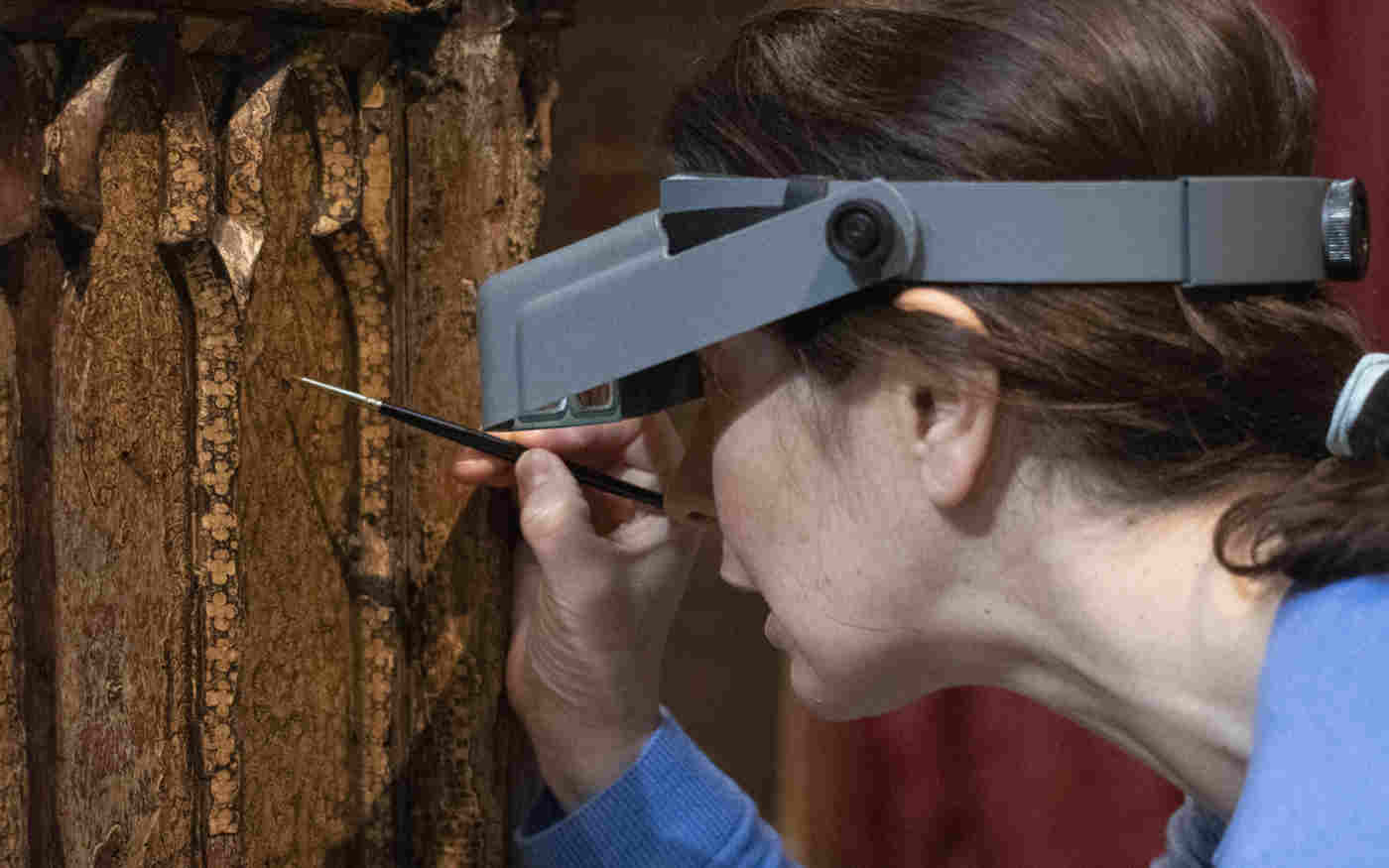 Photograph of woman holding a small paintbrush and wearing a magnifying headset conserving the Coronation Chair