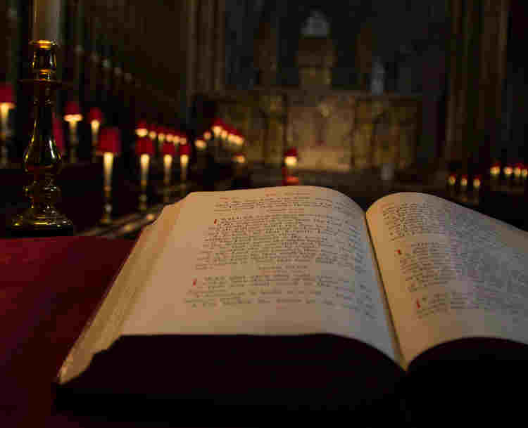 Photograph of an open Bible on a red lectern, in front of a background of lit lamps in the quire of Westminster Abbey