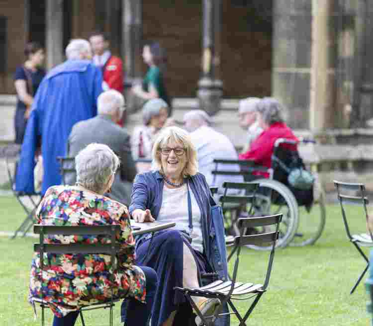 Photograph of two people sitting on chairs talking and enjoying themselves, with other people in the background within Westminster Abbey at an Abbey Association event
