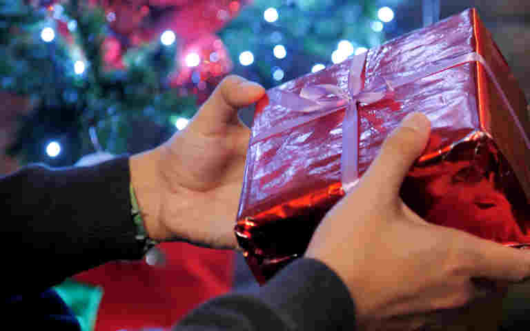 Photograph of hands holding a red Christmas present in front of a Christmas tree with lights