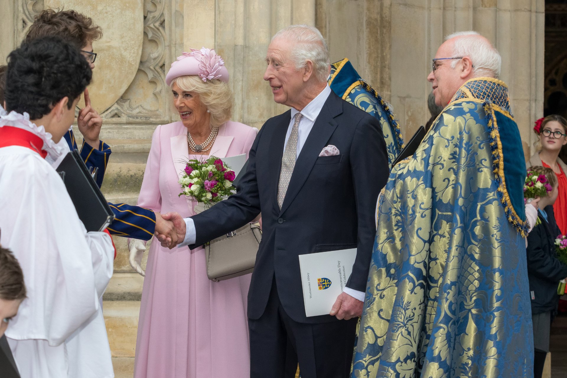King Charles III shaking hands with a chorister. He is standing next to Queen Camilla