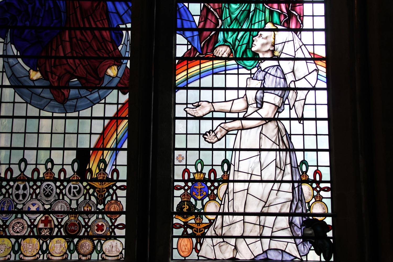 A depiction of a nurse looking up in a stained glass window. A rainbow and feet of other people can be seen behind her.