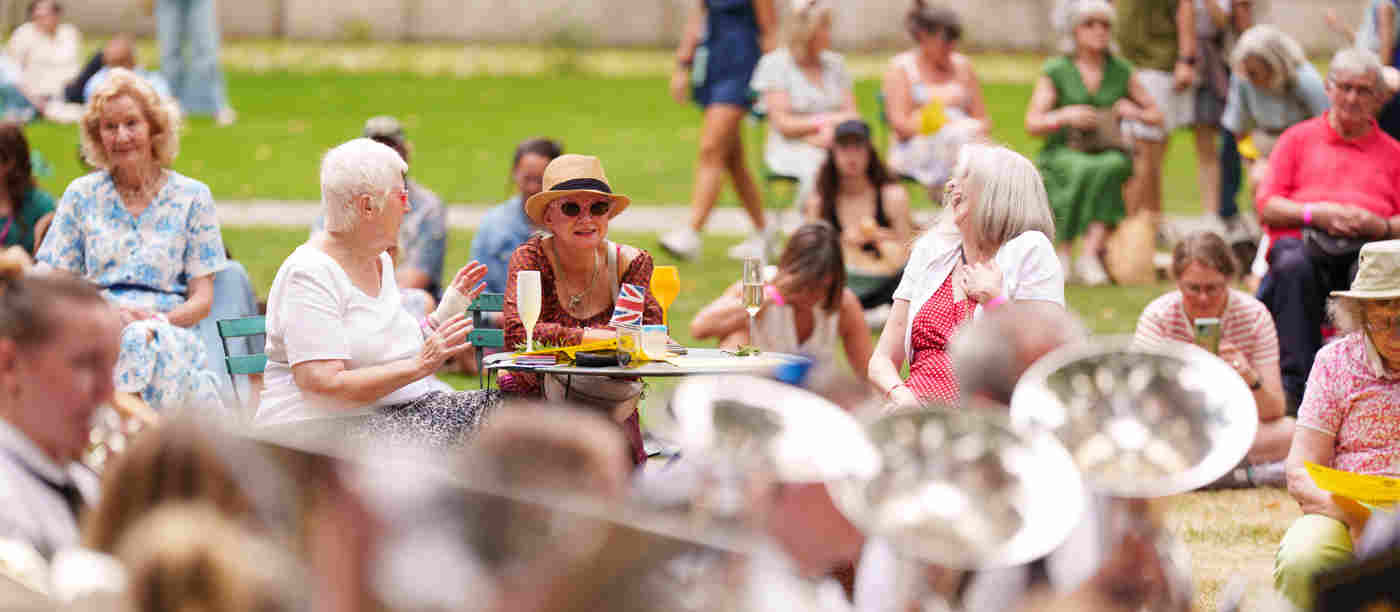 Photograph of visitors chatting, drinking and watching the brass band, in the foreground, for an event in College Garden