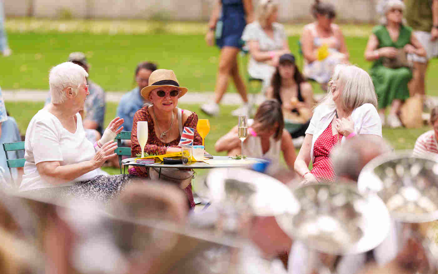 Photograph of visitors chatting, drinking and watching the brass band, in the foreground, for an event in College Garden