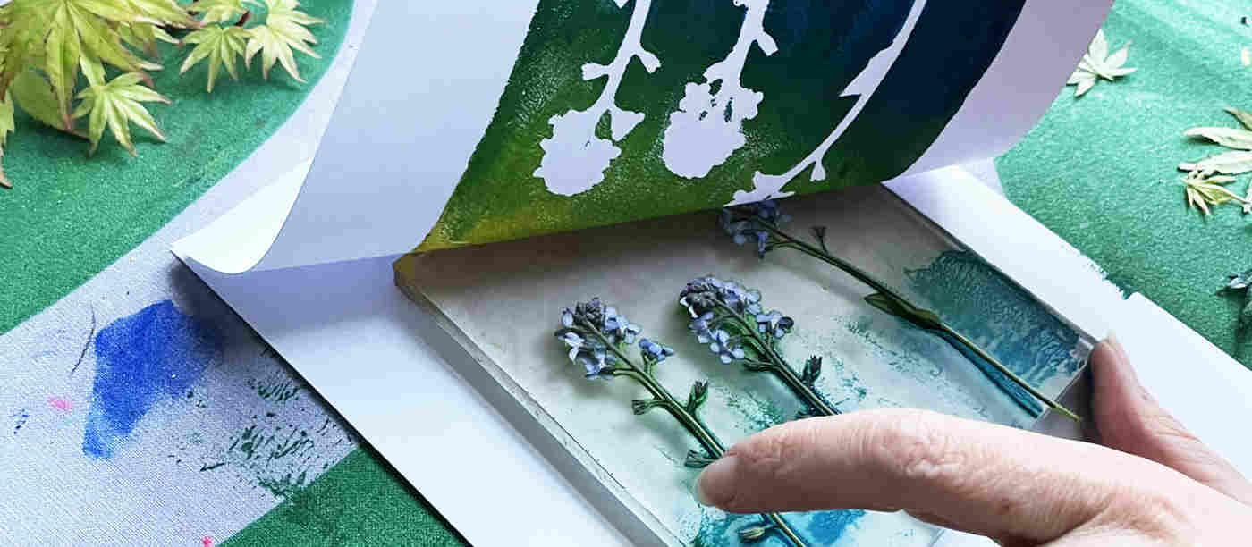 Photograph of gelli printing of plants, with green and blue paint, with a hand holding it in place and another lifting the paper up
