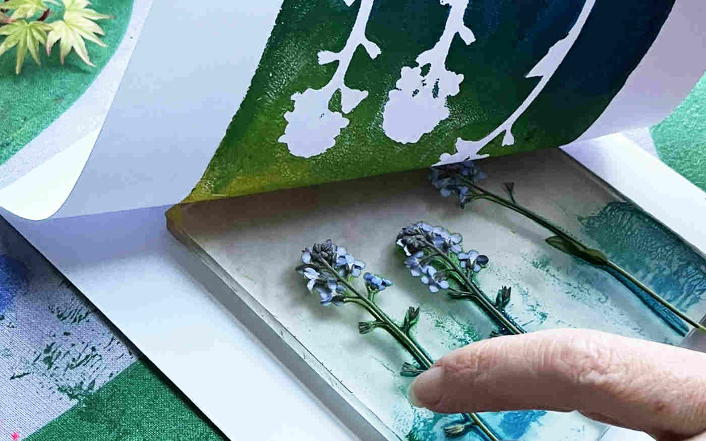 Photograph of gelli printing of plants, with green and blue paint, with a hand holding it in place and another lifting the paper up