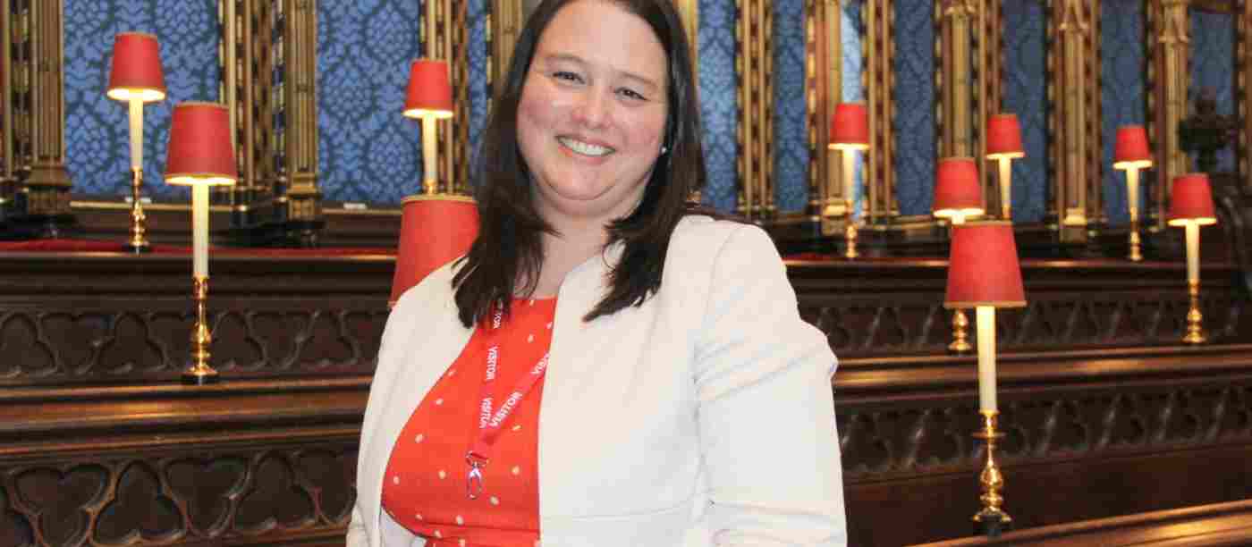 Dr Emma Margrett (a smiling woman who is wearing an orange dress and white jacket) standing in the choir stalls at Westminster Abbey