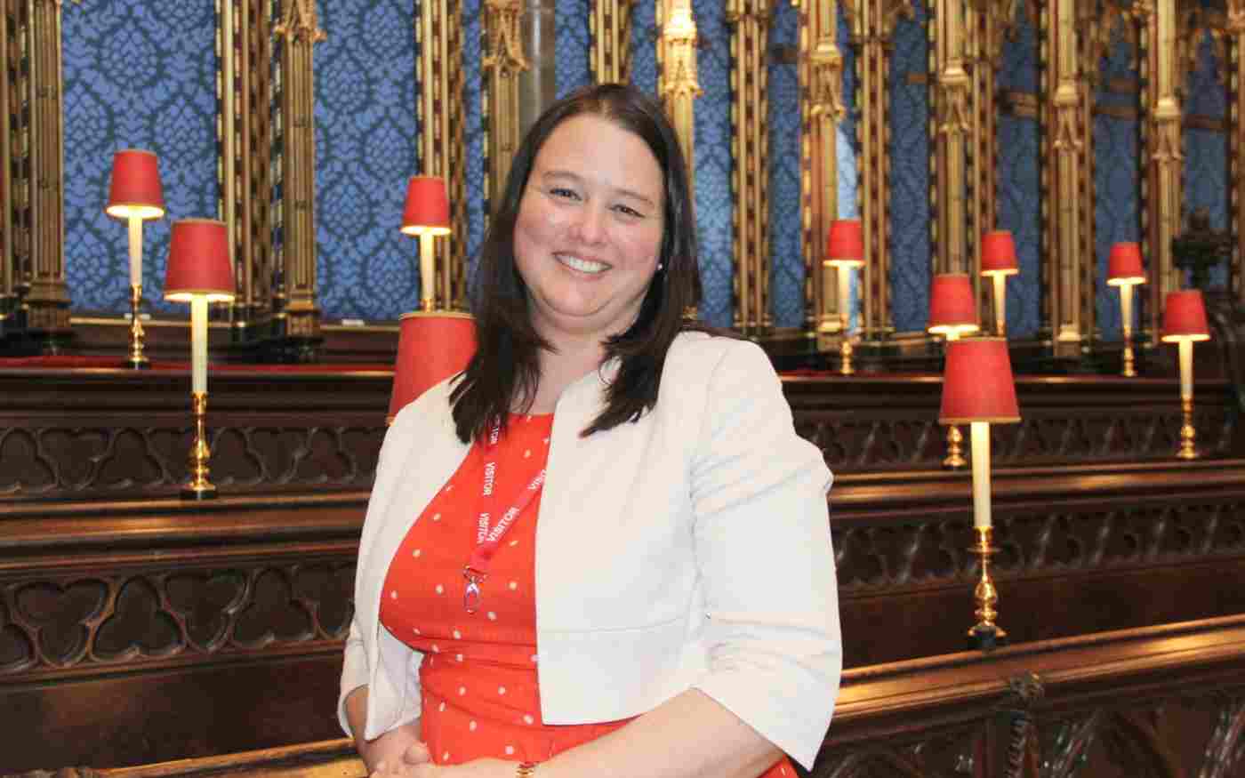 Dr Emma Margrett (a smiling woman who is wearing an orange dress and white jacket) standing in the choir stalls at Westminster Abbey