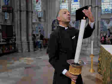 Photograph of male verger wearing a black cassock holding a large church candle and blowing it out within Westminster Abbey