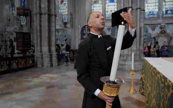 Photograph of male verger wearing a black cassock holding a large church candle and blowing it out within Westminster Abbey