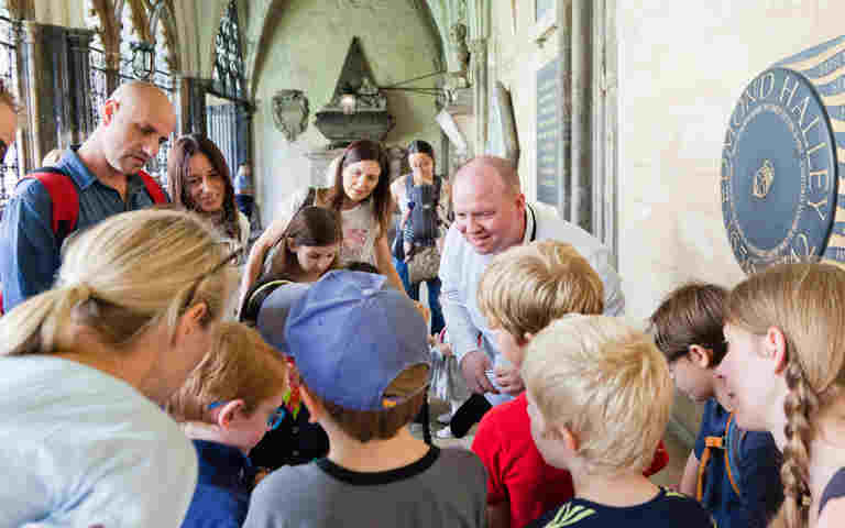 Photograph of an adult leading a tour and listening to feedback from a group of children and their parents within the cloister of Westminster Abbey