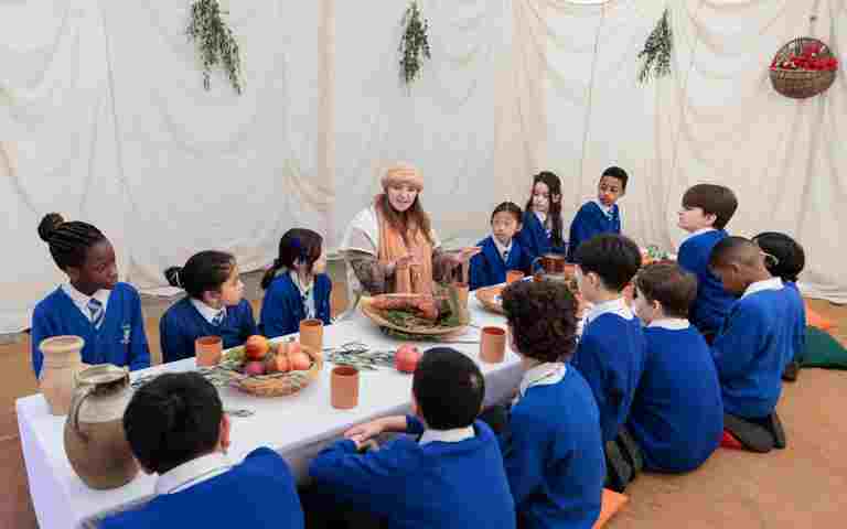Photograph of adult actor surrounded by children dressed in school uniform, sitting on the floor around the low table for a demonstration of the scene of the Last Supper