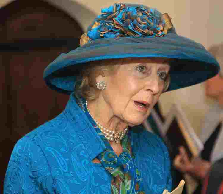 Princess Alexandra wearing a blue hat with blue and gold flowers, a blue dress and pearl necklace