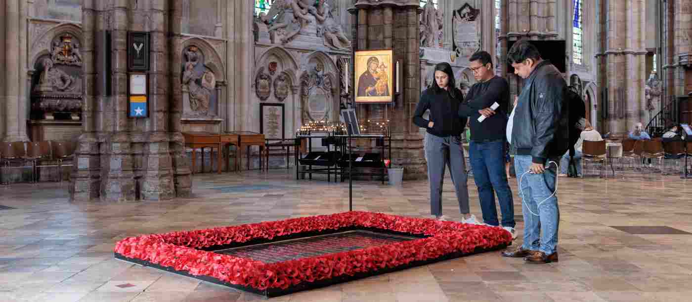 Photograph of a group of three visitors standing by and looking at the Grave of the Unknown Warrior, surrounded by red poppies, in Westminster Abbey