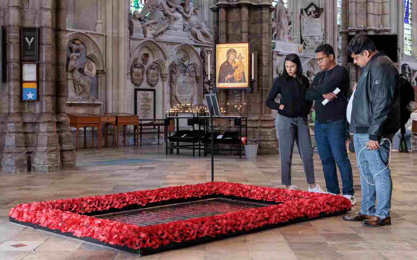 Photograph of a group of three visitors standing by and looking at the Grave of the Unknown Warrior, surrounded by red poppies, in Westminster Abbey