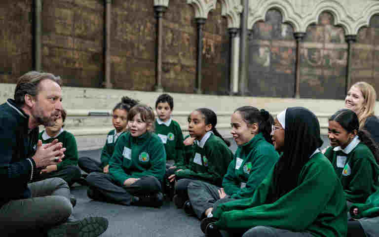 Photograph of a man sitting on the floor speaking to a group of engaged children in school uniform within the Chapter House of Westminster Abbey