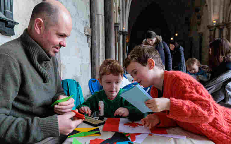 Photograph of a seated adult with two children, leaning over a table covered in paper, glue and other craft activities