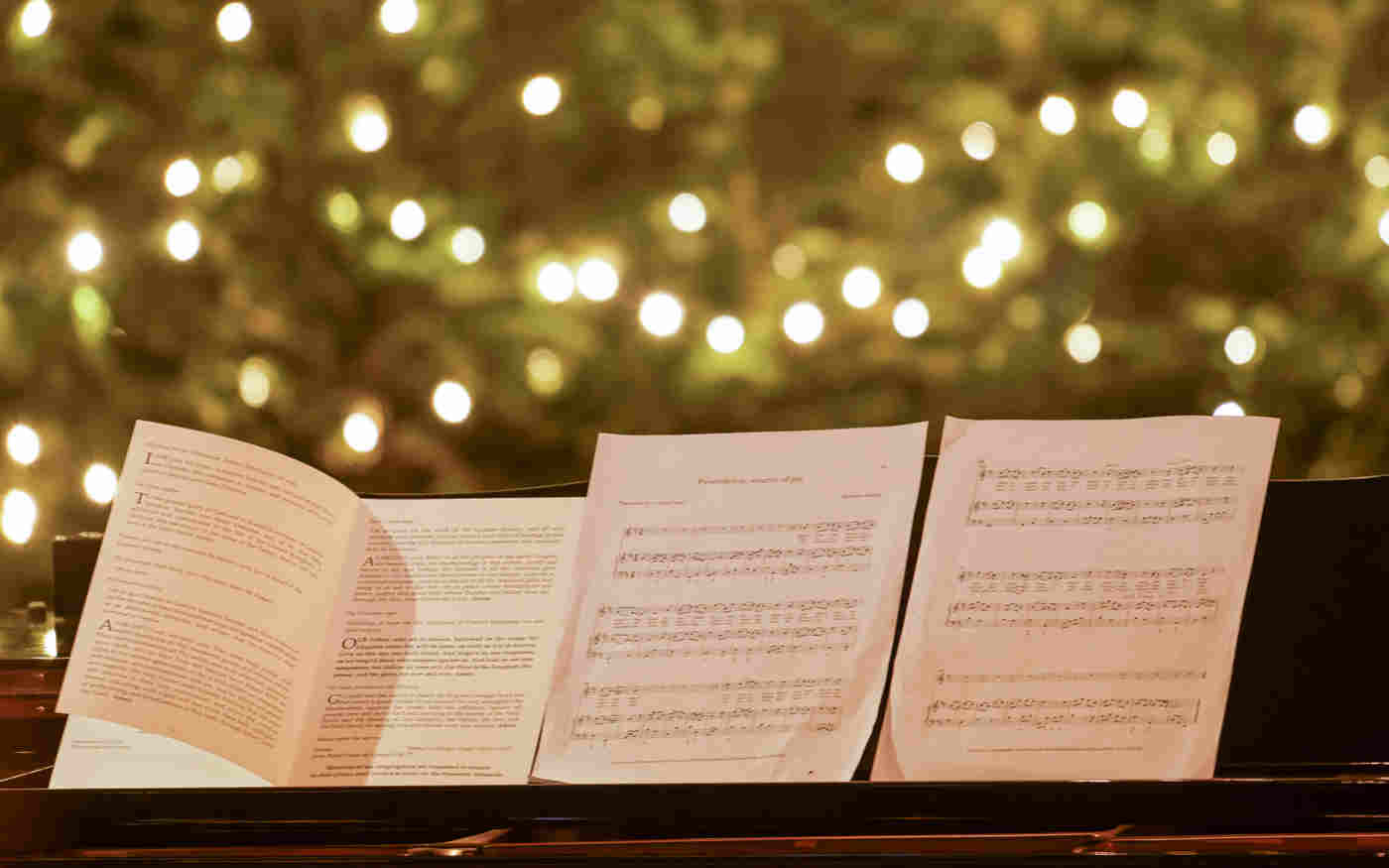 Photograph of a piano with four pages of sheet music and in the background a lit Christmas tree filling the whole space