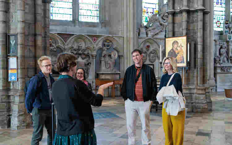 Photograph of a Westminster Abbey member of staff talking to a group of visitors who are smiling, standing within the nave of Westminster Abbey