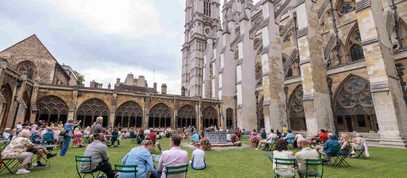 Photograph of groups of visitors sitting on chairs within the garth, an area of grass, with the stone cloisters and West Towers of Westminster Abbey in the background