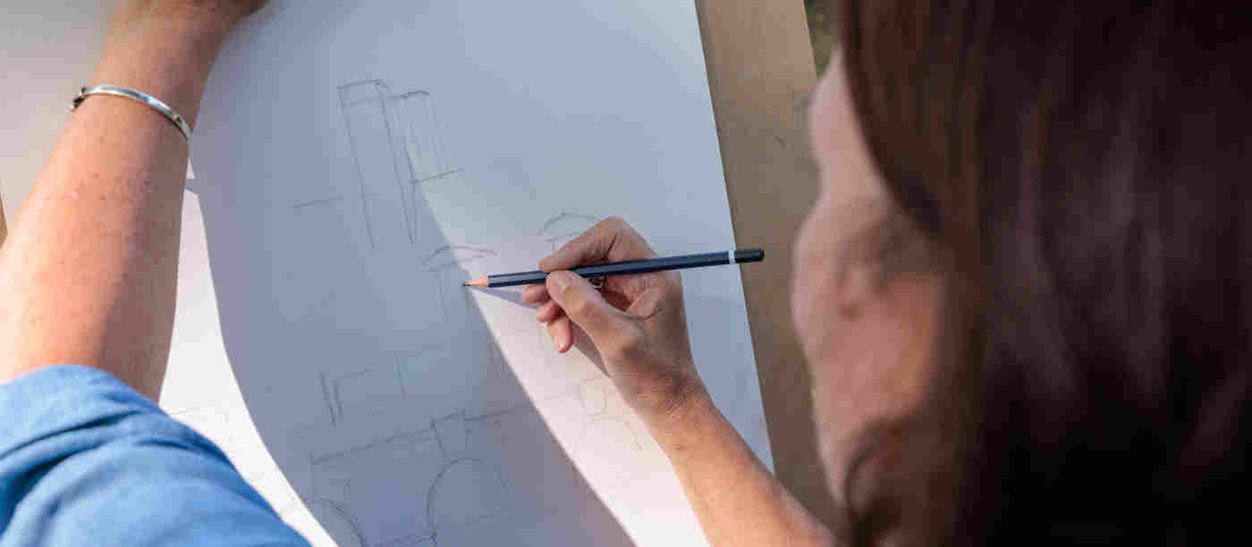 Photograph of the back of a woman's head and arm who is sketching onto a piece of paper on a clipboard