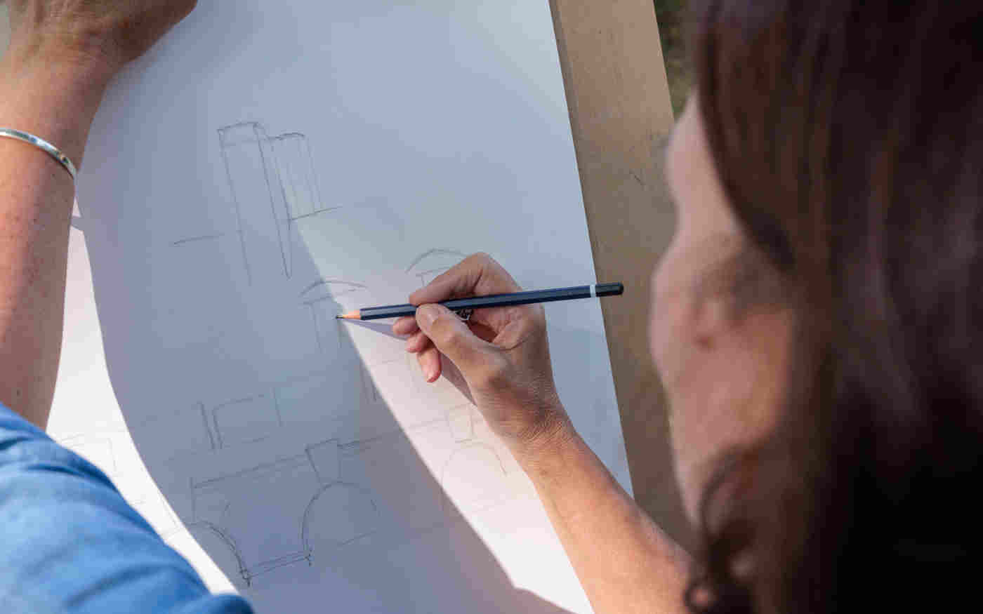 Photograph of the back of a woman's head and arm who is sketching onto a piece of paper on a clipboard
