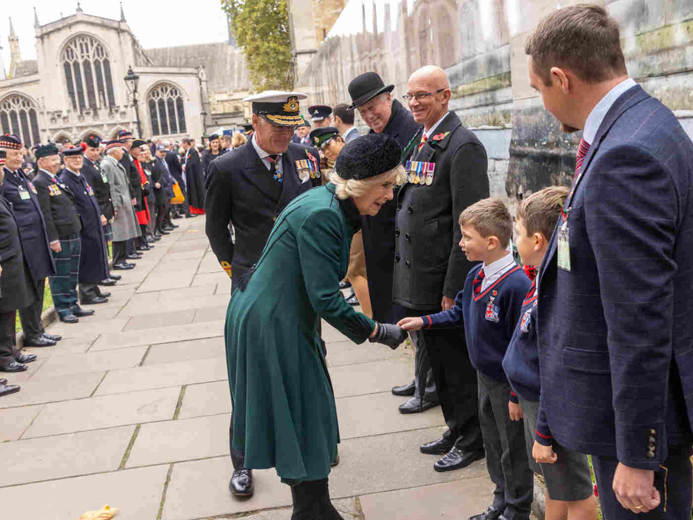 The Queen Consort shaking hands with two schoolchildren. Either side of them are people standing in a line