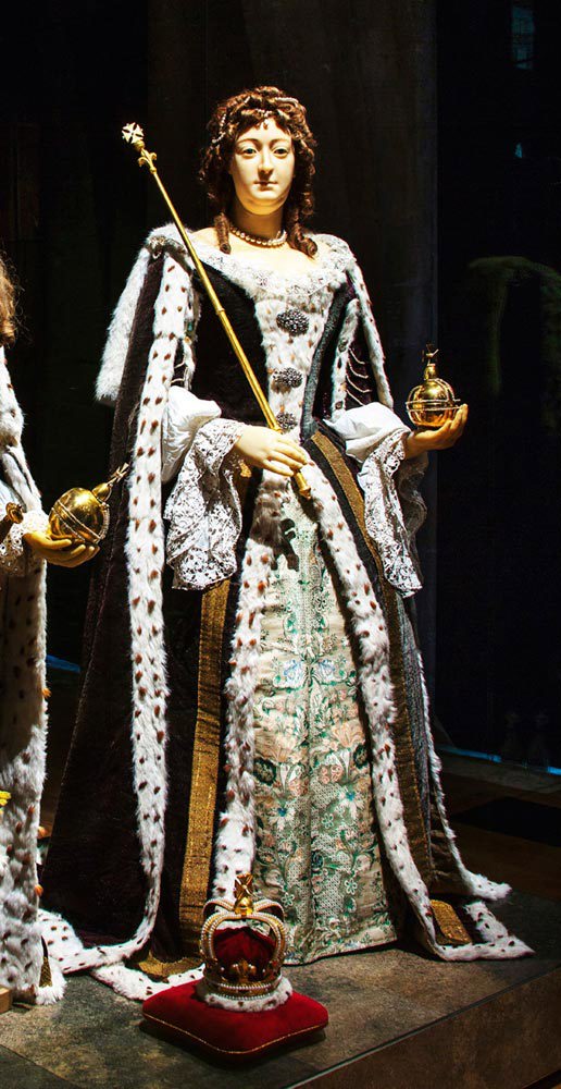Wax effigy of Mary II, wearing elaborate robes, holding a sceptre and orb, with a crown at her feet on a cushion
