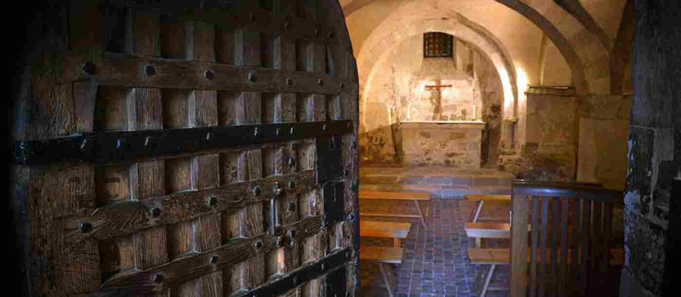A medieval wooden door ajar revealing a small chamber with benches and a stone altar with a crucifix and candles