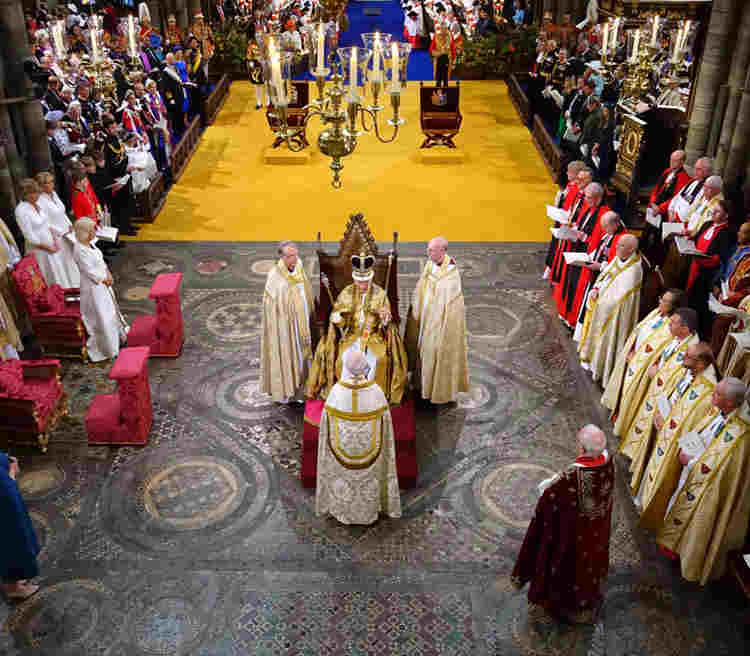 Photograph showing HM King Charles III sitting on the coronation chair within the coronation theatre. The coronation theatre includes a yellow and blue carpet, as well as the uncovered Cosmati Pavement within Westminster Abbey.