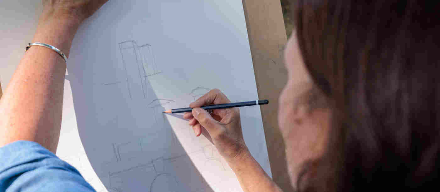Photograph of the back of a woman whose arm is holding a clipboard and with the other hand is sketching with pencil an image