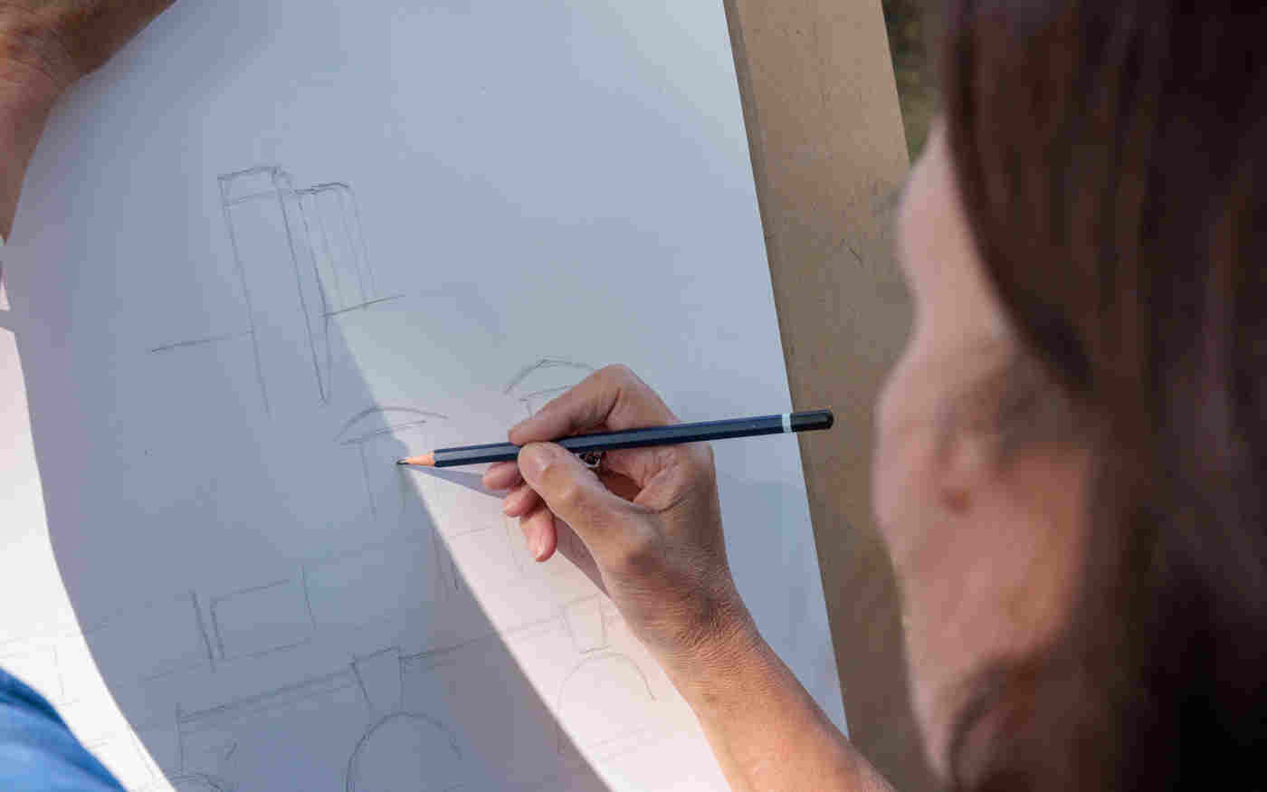 Photograph of the back of a woman whose arm is holding a clipboard and with the other hand is sketching with pencil an image