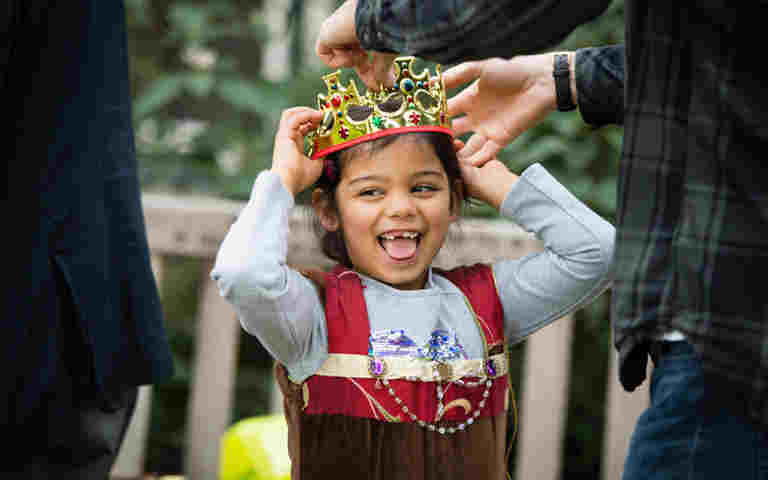 Photograph of young child smiling while a fake crown is being placed on her head by an adult, representing What happens in a coronationactivity pack