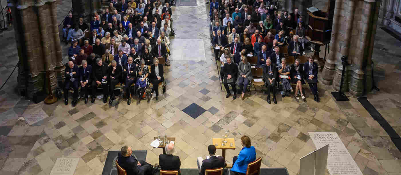 Photograph looking down on a panel of speakers from behind and seeing the front of audience members in the nave of Westminster Abbey