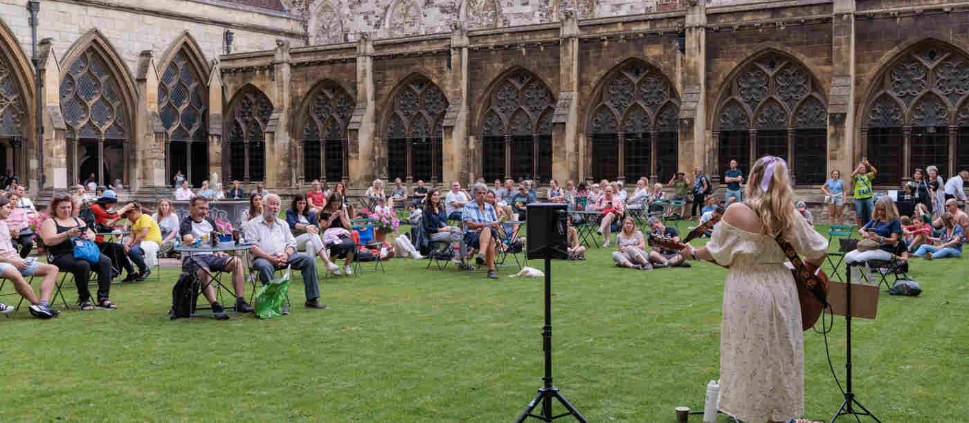 Photograph of visitors in the cloister garth, an area of grass, within Westminster Abbey, listening to a musician playing the guitar