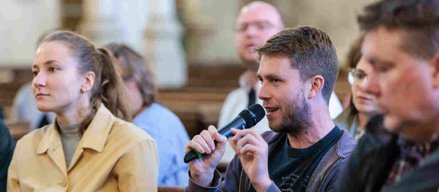 Photograph of an audience member holding a microphone asking a question surrounded by other people in St Margaret's Church